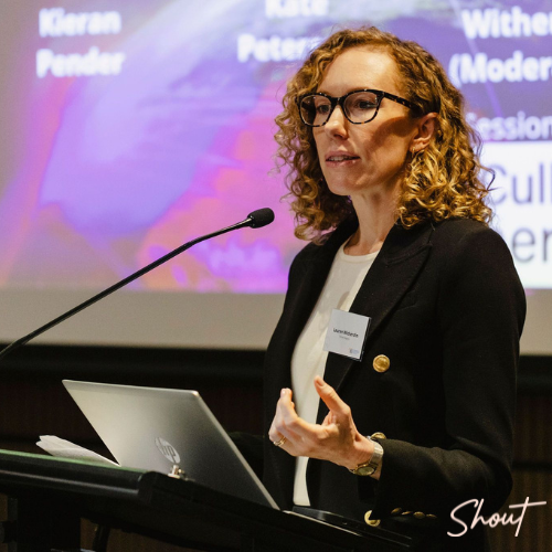 A woman with curly hair speaks at a podium during a professional event.