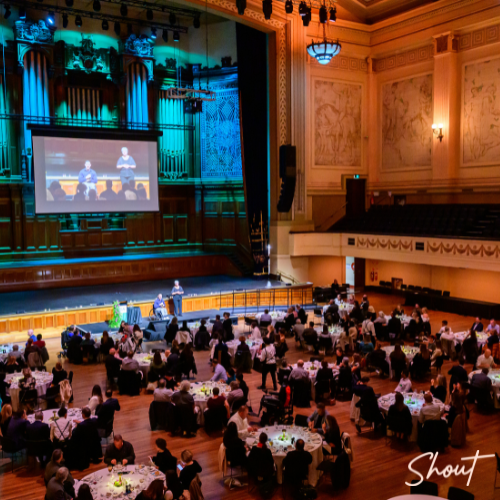 Audience watching a presentation in an ornate theater with round tables.