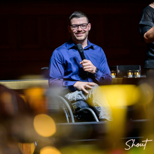 Man in wheelchair speaking on stage with microphone.