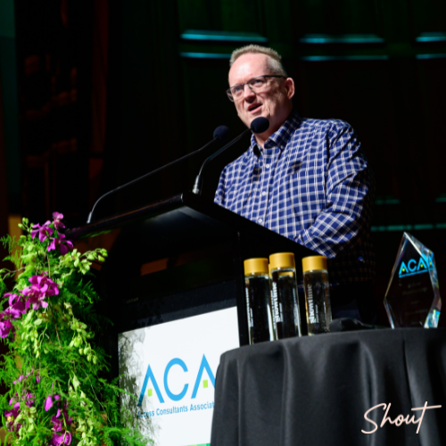 Man speaking at a podium with awards on a table nearby.