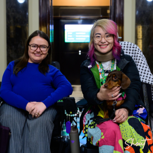 Two women smiling, one holding a dog, both in wheelchairs.
