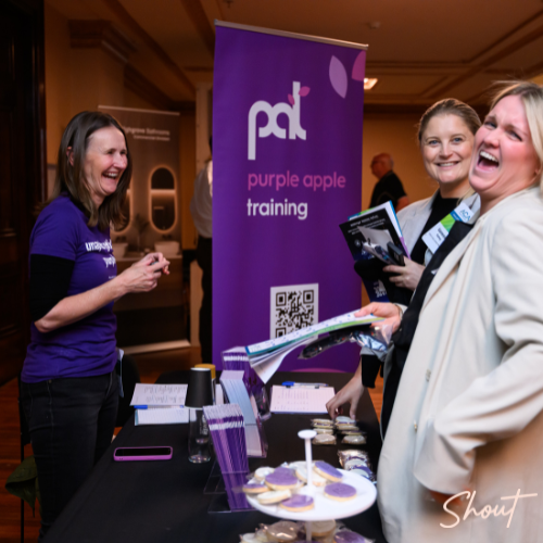 A woman explains Purple Eagle Training at a booth with attendees listening.