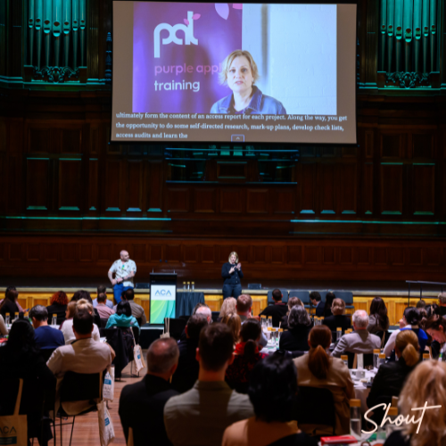 Audience applauds a speaker at a conference in a grand hall.