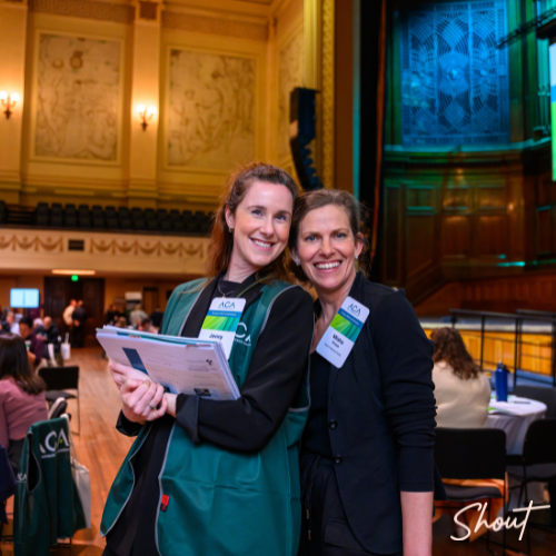 Two women smiling at an indoor event, one holding a booklet.